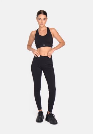 Fekete sportos toppot, magas derekú leggingset és fekete tornacipőt viselő nő áll egy fehér háttér előtt.