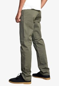 Olivgrüne Baumwollchinos mit geradem Bein, ausgestattet mit seitlichen Taschen und sichtbaren Nähten, kombiniert mit schwarzen Schuhen und Gummisohlen.