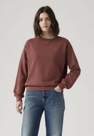 Frau steht mit der Hand in der Tasche, trägt ein dunkelrotes Sweatshirt und blaue Jeans vor schlichtem hellem Hintergrund.