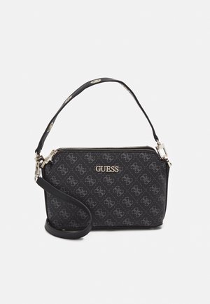 Pochette noire de la marque Guess avec motif logo intégral, garniture dorée, fermeture éclair et bracelet de poignet amovible sur fond blanc.