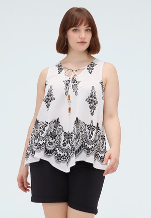 Kvinde iført hvid ærmeløs top med sorte blomsterprint og sorte knælange shorts, stående mod en ensfarvet baggrund.