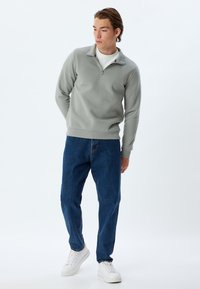 Felpa grigia chiara con zip e colletto alto, abbinata a jeans in denim blu scuro a taglio largo e sneakers bianche. Texture complessiva liscia.