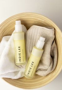 Dwie żółte butelki oznaczone "INDIE LEE SOOTHING CLEANSER" znajdują się w bambusowej misce, w towarzystwie białych i teksturowanych beżowych ręczników.
