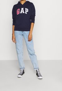 Felpa navy con logo "GAP" in rosso e grigio, abbinata a jeans azzurri chiari e sneakers nere alte. Look casual e vestibilità rilassata.