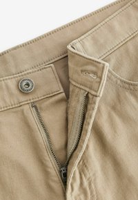 Taille de pantalon beige avec fermeture éclair métallique ouverte, rabat à boutonnière et bouton en métal gravé « Premium Denim Goods Since 1989 ».