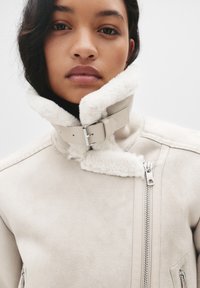 Beżowa skórzana kurtka z wysokim kołnierzem z owczej wełny, metalowym zapięciem na klamrę i asymetrycznymi zamkami z przodu. Miękka tekstura i dopasowany krój.