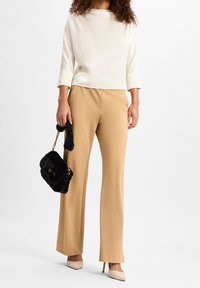 Haut à manches trois-quarts couleur crème, pantalon large beige, talons couleur chair, et sac à main en fausse fourrure noire avec une bandoulière chaîne dorée.