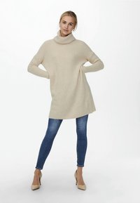 Beige oversized gebreide trui met een hoge kraag en geribbelde textuur, gecombineerd met strakke blauwe jeans en lichtgekleurde hakken.