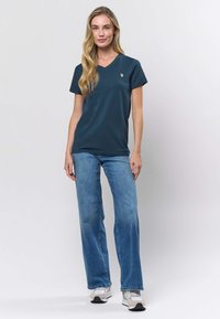 Tirkīza krāsas T-krekls ar V-veida kakla izgriezumu un īsām piedurknēm, ar nelielu zelta logotipu. Sabalansēts ar gaiši zilām, platām džinsu biksēm un pelēkām sporta apaviem.