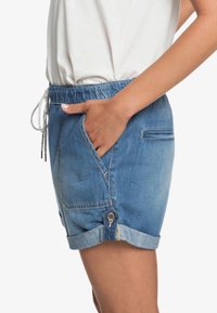 Short en denim avec un lavage bleu clair, une ceinture élastique avec cordon, et des poches latérales. Ourlet retroussé et détail de fermeture par bouton.