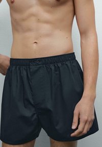 Shorts noirs avec une taille élastique et une fermeture à bouton, fabriqués en tissu lisse, présentent un design simple et une coupe ajustée.