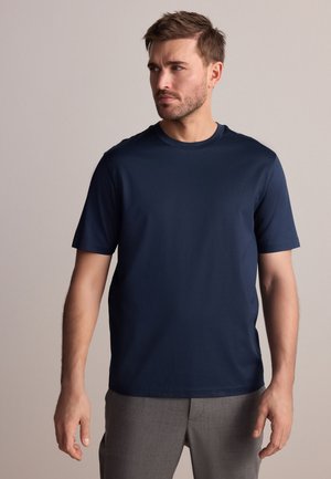 Mand med kort brunt hår iført en ensfarvet navyblå kortærmet skjorte og grå bukser, der kigger mod venstre mod en neutral baggrund.