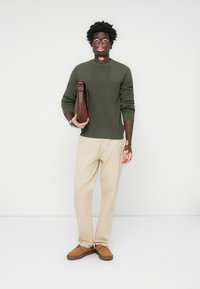 Olive green ribbed sweater, beige chinos, and tan shoes. Holding a brown leather bag, posed against a plain white background.