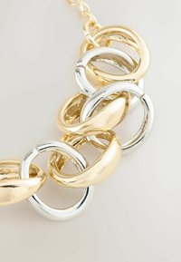 Catene spesse con maglie alternate in tonalità d'oro e argento, con una finitura lucida e brillante su uno sfondo chiaro.