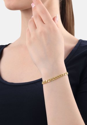 Eine Frau trägt ein Armband aus goldenen Kettengliedern an ihrem Handgelenk, während ihre Finger sanft ihr Gesicht berühren. Sie ist in einem schwarzen Oberteil gekleidet.