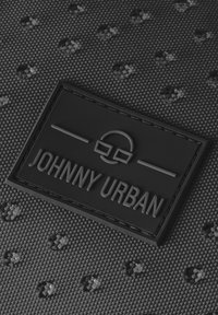 Mustikka teksturoitu kangas, jossa on kohotettu kumilappu, jossa on präntätty "JOHNNY URBAN" ja graafinen suunnittelu. Sisältää pieniä pistekuvioita kauttaaltaan.