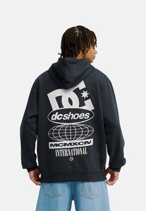 Osoba ubrana w czarną bluzę z kapturem z dużymi białymi logotypami i napisami DC Shoes, w tym grafiką globusa, widoczna od tyłu. Widoczne niebieskie dżinsy.