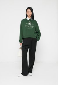 Felpa verde con colletto bianco e logo grafico, abbinata a pantaloni neri a gamba larga e sneakers bianche. Il modello tiene in mano una piccola borsa bianca.