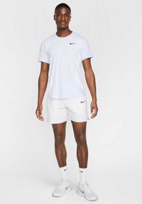 Gaiši zilas īsām piedurknēm Nike T-krekls ar melnu logo, apvienots ar baltām šortiem, baltām zeķēm un gaišām sporta kurpēm. Gluda auduma faktūra.