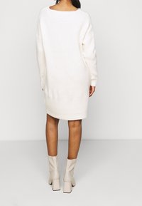 Sweter sukienkowy w kolorze kremowym, o luźnym kroju, prążkowanej teksturze i wycięciu na plecach. W zestawie z jasnobeżowymi botkami na blokowym obcasie.