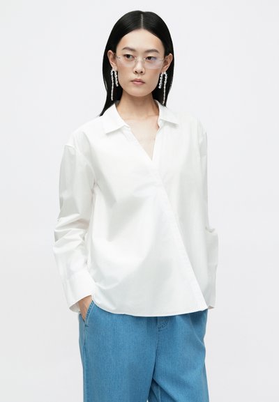 Camisa branca de manga longa, com colarinho e um design de envoltório assimétrico, combinada com calças de ganga azul claro. Textura suave, estilo minimalista.