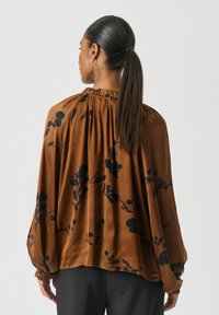 Brun blus med svarta blommönster, med en samlad halsringning och utsvängda ärmar, tillverkad av lättviktigt tyg.