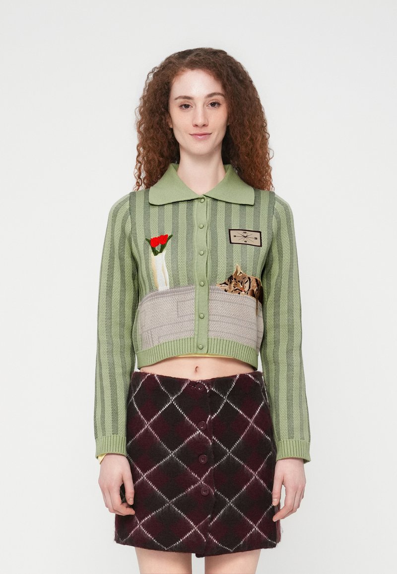 Πράσινη κοντή ζακέτα με κάθετες ρίγες, κεντητά λουλούδια και μοτίβα γάτας· συνδυασμένη με μαύρη και μπορντό φούστα σε σχέδιο αργκόιλ με κουμπιά.