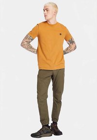 T-shirt orange à manches courtes, coupe slim, avec un petit logo sur la poitrine. Pantalon de jogging vert olive avec poignets élastiques et chaussures de randonnée noires.