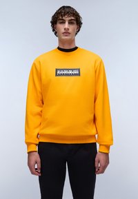 Heldere gele trui met een geribde halslijn en manchetten, voorzien van een zwart rechthoekig logo op de borst. Zwarte broek maakt de look compleet.
