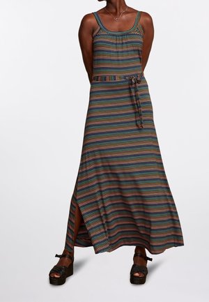 Vrouw in een mouwloze maxi-jurk met horizontale strepen in meerdere kleuren, voorzien van een zijsplit en bijpassende riem, gecombineerd met zwarte platformsandalen.