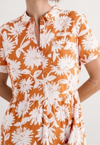 Orange button-up dress with short sleeves, featuring large white floral patterns, a chest pocket, and a gathered waist for a relaxed fit.