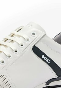 Zapatillas de cuero blancas con una franja de acento negra, detalles perforados, cordones redondos y un logotipo cerca de los ojales.