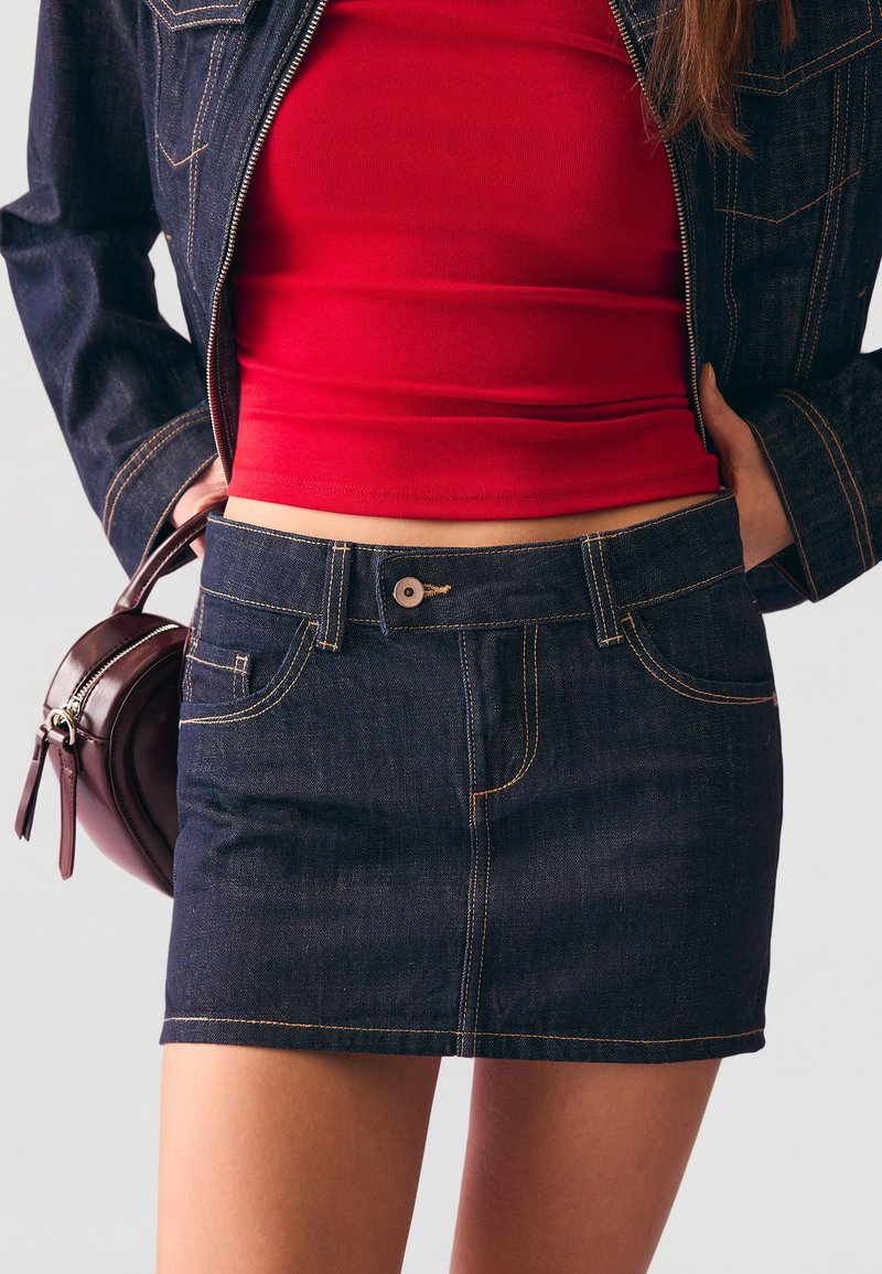 Femme portant une jupe en denim foncé et une veste avec un crop top rouge, tenant un petit sac à main marron foncé à ses côtés.