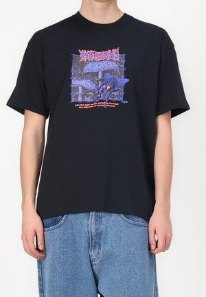 Persona che indossa una maglietta nera con grafica di insetti e funghi blu e viola e testo arancione, abbinata a jeans blu larghi.