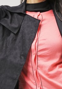 Svart mockajacka över en rosa knappad cardigan med tunna svarta sömmar längs knapplinjen. Hög halsstil.