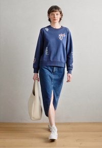 Sweatshirt bleu avec des accents brodés visibles, associé à une jupe en denim avec une fente devant. Un sac fourre-tout neutre et des baskets blanches complètent le look.