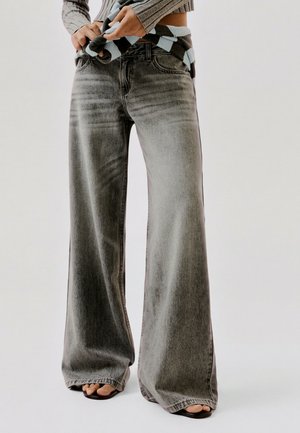 Personne portant un jean gris délavé à jambes larges, des sandales ouvertes et un pull court, les mains ajustant un objet à motifs à la taille.