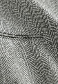 Gros plan sur un tissu tweed chevron gris et blanc avec un détail de poche passepoilée cousue.