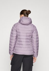 Lys lilla pufferjakke med hette, som har horisontal søm og en glatt, skinnende tekstur. Båret over svarte bukser.