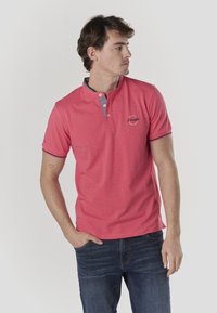 Rosa Kurzarm-Poloshirt mit einem Knopfkragen, verziert mit kleinen blauen und weißen Mustern, und einem Logo auf der linken Brustseite. Kombiniert mit blauen Jeans.