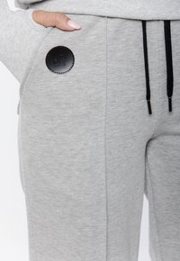 Szare dresowe spodnie z miękkiego materiału z czarną skórzaną naszywką z logo. Widoczne są regulowane czarne sznurki oraz boczne kieszenie.