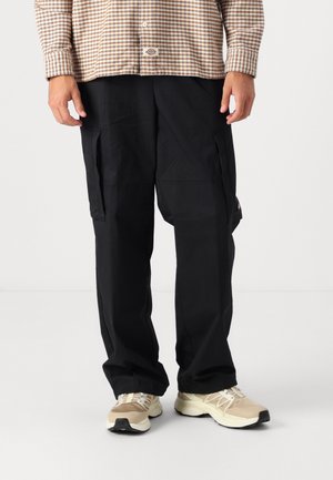 Person iført sorte cargobukser, beige sneakers og en brunternet skjorte stående mod en ensfarvet baggrund.