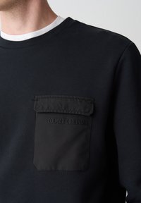 Man draagt een zwart sweatshirt met een zwart klepzakje op de borst met het label "COLMAR ORIGINALS" boven een witte ondershirt met ronde hals.