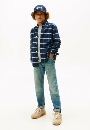 Niño con cabello rizado que lleva una gorra azul marino, una camisa de cuadros en tonos azul marino, una camiseta gris, jeans doblados y zapatillas beige, de pie con una mano en el bolsillo.