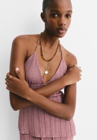 Halenka ve dusty rose barvě na halter, s texturovanou tkaninou, hlubokým výstřihem do V, vrstveným korálkovým náhrdelníkem a stříbrnými akcenty. Modelka má založené ruce na prsou na jednobarevném pozadí.