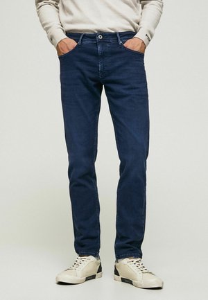 Person trägt dunkelblaue, eng anliegende Jeans und weiße Turnschuhe, steht mit den Händen in den Taschen vor einem schlichten Hintergrund.