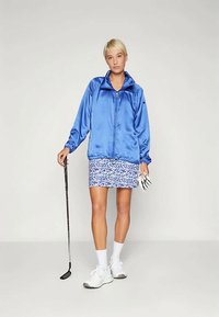 Mėlynas satino užtrauktukas su aukštu apykakle, dėvimas ant mėlynos ir baltos leopardų rašto sijono. Balti sportiniai bateliai ir golfo lazda rankoje.