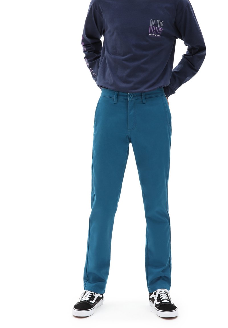 Homme portant un pantalon slim turquoise, des baskets noires et blanches, et un t-shirt Vans bleu marine à manches longues, debout les mains derrière le dos.