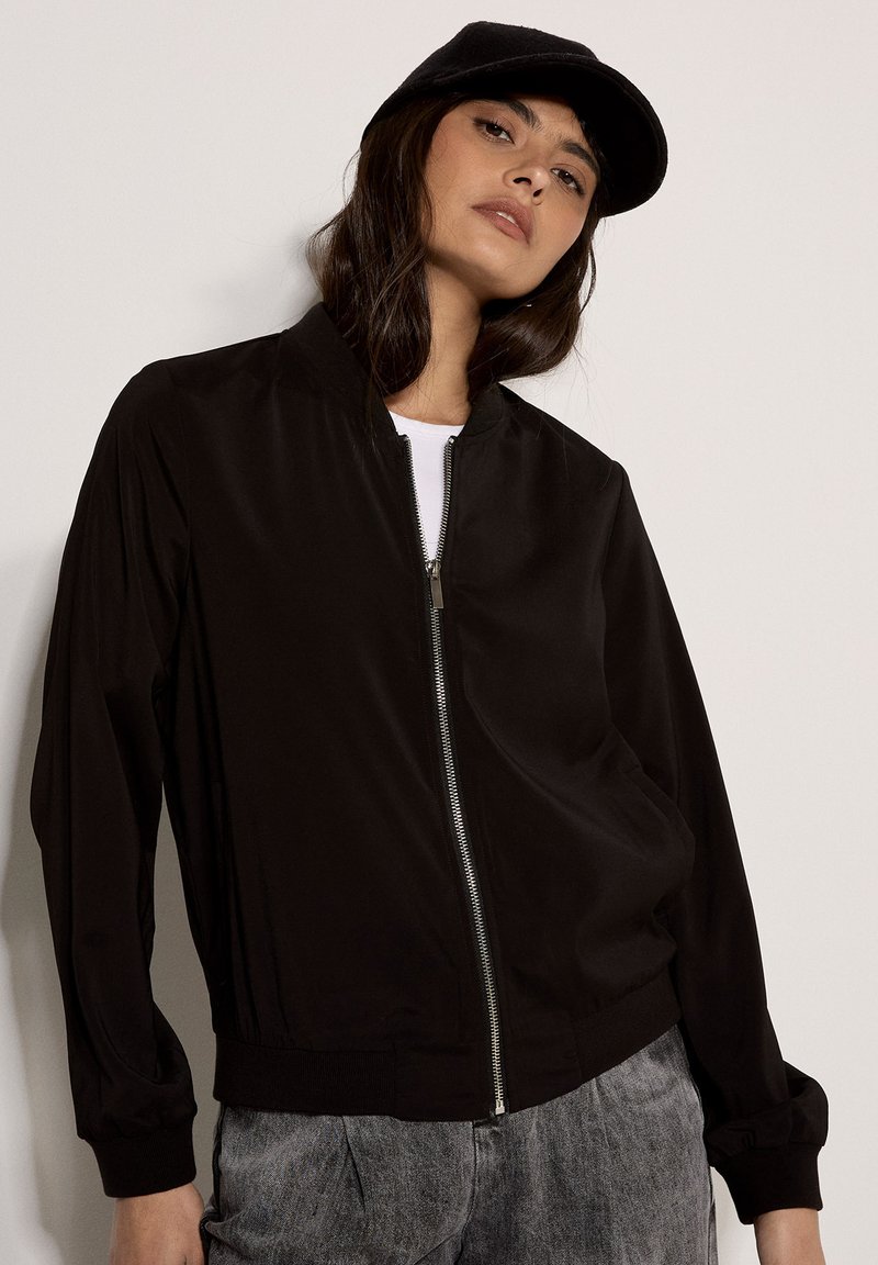 Femme portant une veste bomber noire zippée, une casquette noire, un haut blanc et un pantalon gris, debout devant un fond clair uni.