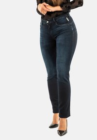 Jeans skinny de mezclilla azul oscuro con una textura suave, diseño de cinco bolsillos y detalles en hardware plateado, combinados con zapatos de tacón alto negros.